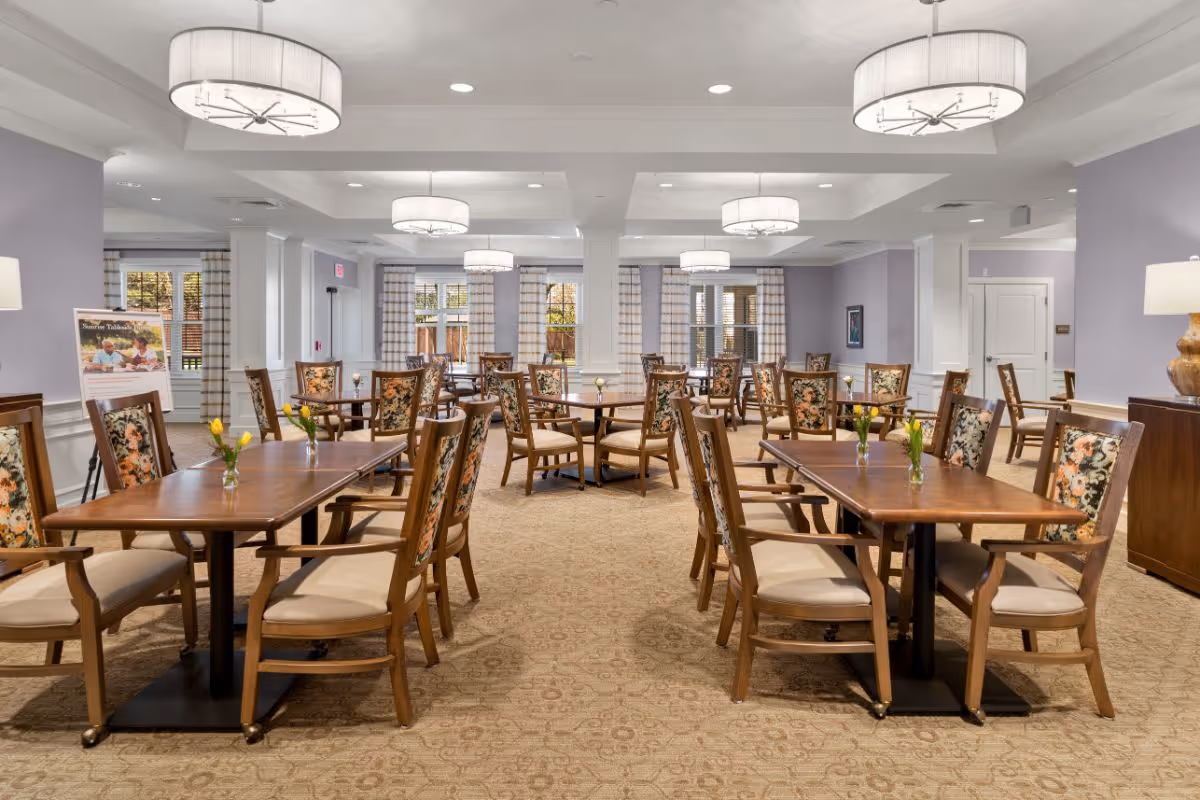 A spacious dining room with multiple wooden tables and chairs featuring floral upholstery. Each table has a small vase with yellow flowers. The room has soft purple walls, large windows with striped curtains, and modern ceiling lights. There is a sign on an easel in the back left corner and a wooden sideboard with a lamp on the right.