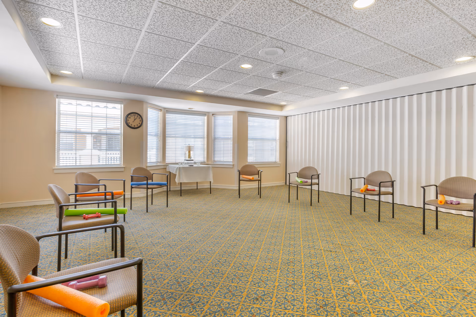 A spacious room with patterned carpet and beige walls, featuring a circle of chairs each holding colorful exercise equipment like foam rollers and dumbbells. Large windows with blinds let in natural light, and a clock is mounted on the wall near the windows. A table covered with a white cloth is positioned in front of the windows.