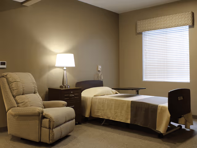 A simple, clean bedroom in a senior living facility featuring a single bed with beige and brown bedding, a beige recliner chair, a dark wooden nightstand with a lamp, and a window with blinds and a patterned valance.
