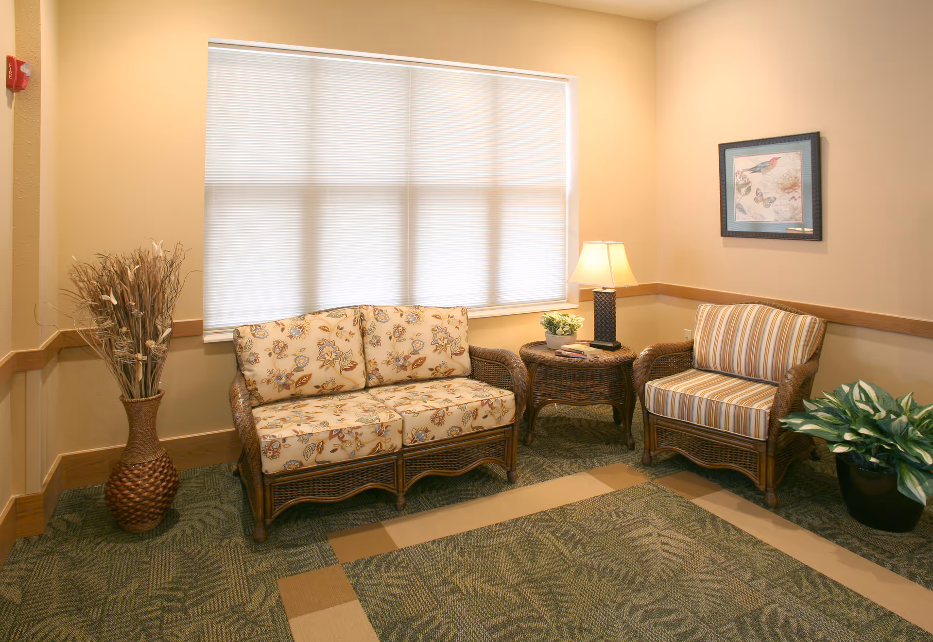A cozy sitting area with a floral-patterned loveseat and a striped armchair made of wicker. Between them is a wicker side table with a lamp and a small plant. There is a large window with closed blinds behind the loveseat, a framed picture on the wall, a tall vase with dried decorative branches on the left, and a potted green plant on the right. The room has beige walls and a patterned green carpet.
