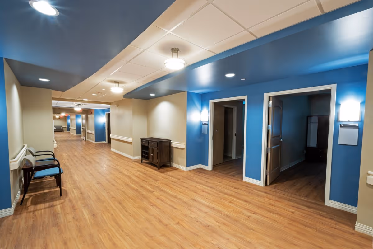 A wide hallway in a senior living facility with wood flooring, beige and blue walls, ceiling lights, and a few chairs along the left wall. Two open doorways on the right lead to rooms with wooden furniture inside.
