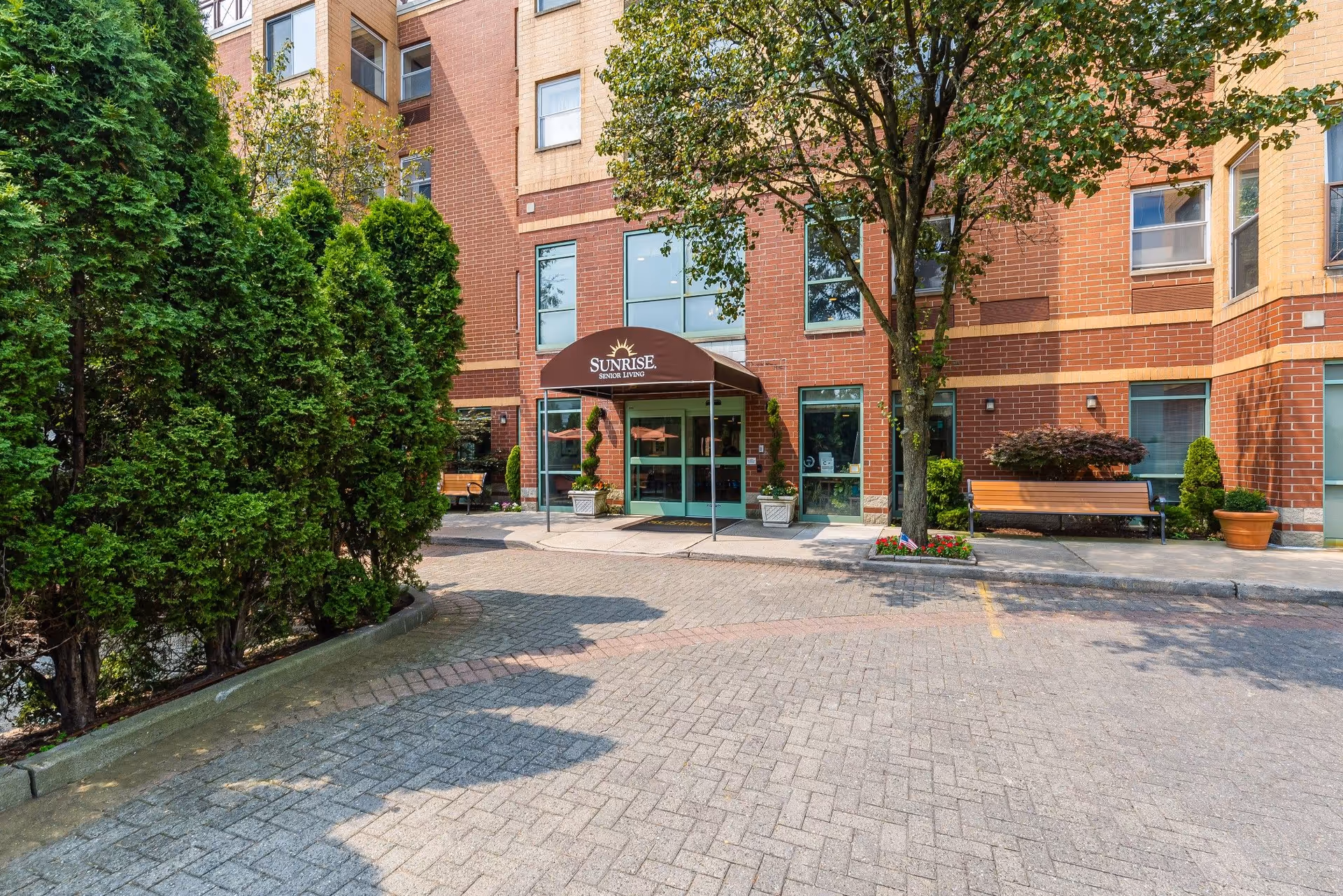 Entrance to Sunrise Senior Living facility with a brown awning displaying the Sunrise logo. The building is made of red brick with several windows. There are green trees and shrubs on the left side and a tree with a bench and potted plants on the right side near the entrance.