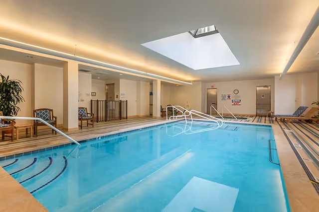 Indoor swimming pool with clear blue water, surrounded by striped flooring and several chairs along the walls. The ceiling features a large skylight allowing natural light to illuminate the area.