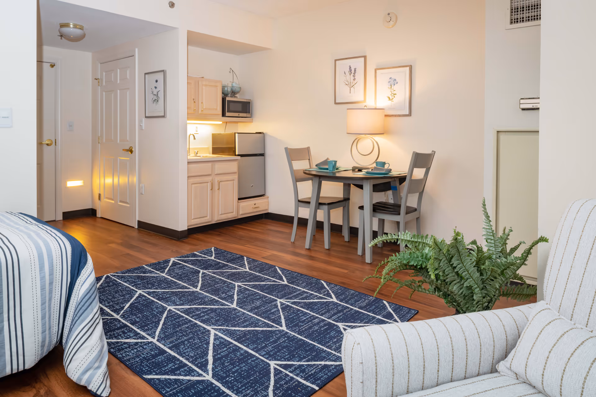 A cozy senior living apartment interior featuring a small kitchenette with light wood cabinets, a mini refrigerator, and a microwave. A round dining table with two chairs is set with teal cups and plates, illuminated by a table lamp. The room has wood flooring with a blue geometric patterned rug, a striped armchair, and framed botanical prints on the wall.