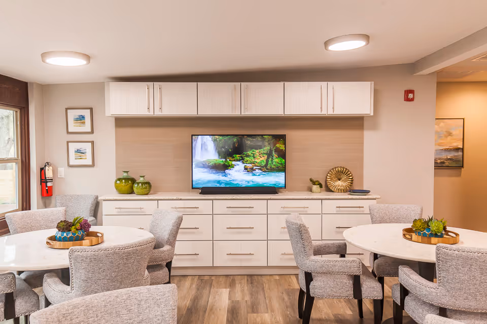 A bright and modern common area with two round white tables surrounded by gray upholstered chairs. A flat-screen TV displaying a nature scene with a waterfall is mounted on a beige wall above a long white cabinet with drawers. Decorative green vases and small plants are placed on the cabinet. The room has wood flooring, recessed ceiling lights, and framed artwork on the walls.