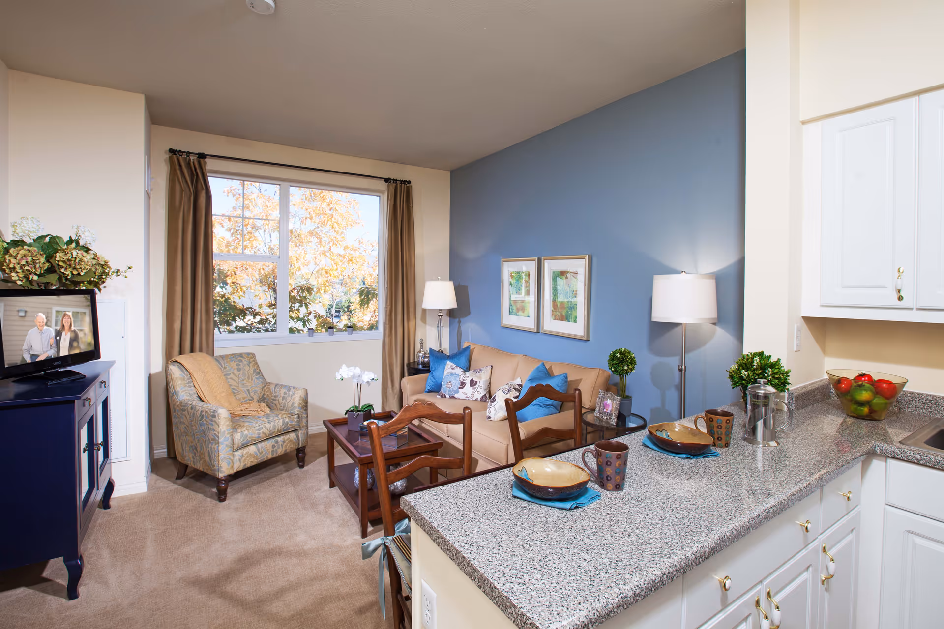 A cozy living area in a senior living facility featuring a beige sofa with blue and patterned pillows, a patterned armchair, a wooden coffee table, and a window with brown curtains. The room has a blue accent wall with two framed pictures and two floor lamps. In the foreground, there is a granite countertop with two place settings including bowls, plates, and mugs. White kitchen cabinets and a bowl of fruit are visible on the right side.