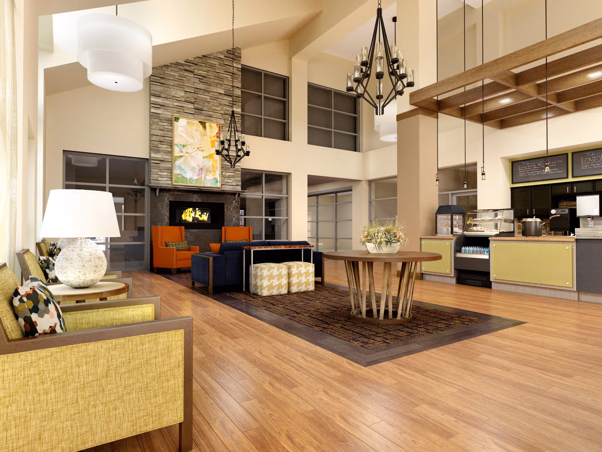 A spacious and well-lit common area with wooden flooring, featuring a seating area with yellow and orange armchairs around a stone fireplace. There is a round wooden table with a floral arrangement in the center on a patterned rug. To the right, there is a counter area with a coffee machine, display case, and menu boards above. Large windows and modern light fixtures hang from the high ceiling.