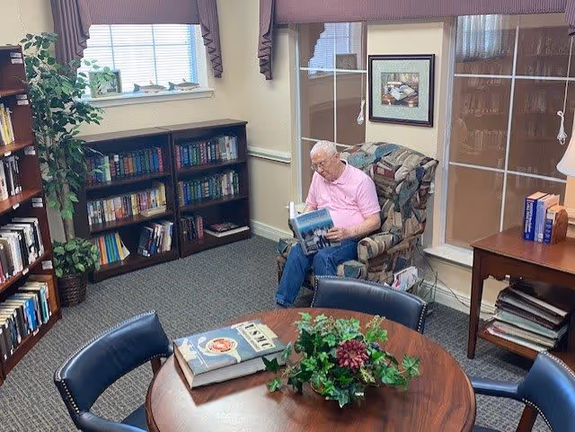 A senior man wearing a pink shirt and blue pants is sitting in a patterned armchair reading a book in a cozy room. The room has bookshelves filled with books, a round wooden table with a plant centerpiece and books on it, and a window with blinds and valances. There is also a side table with books and a lamp.