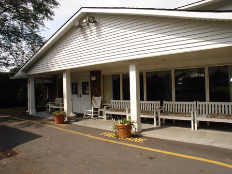 Exterior view of a building entrance with a covered porch area featuring several white wooden benches with cushions and potted plants. The building has white siding and a double door entrance. There are trees and greenery visible in the background.