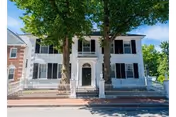 Front exterior view of a two-story white house with black shutters, a black front door, and a white picket fence. Two large trees are in front of the house, and there is a sidewalk and street in the foreground.