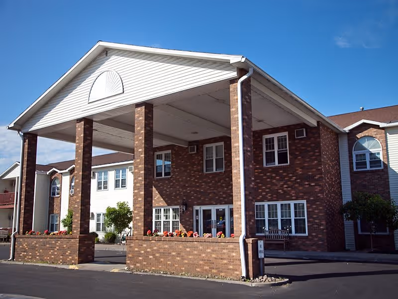 Exterior view of a senior living facility building with a large covered entrance supported by four brick columns. The building has a combination of brick and light-colored siding, multiple windows, and a clear blue sky in the background.
