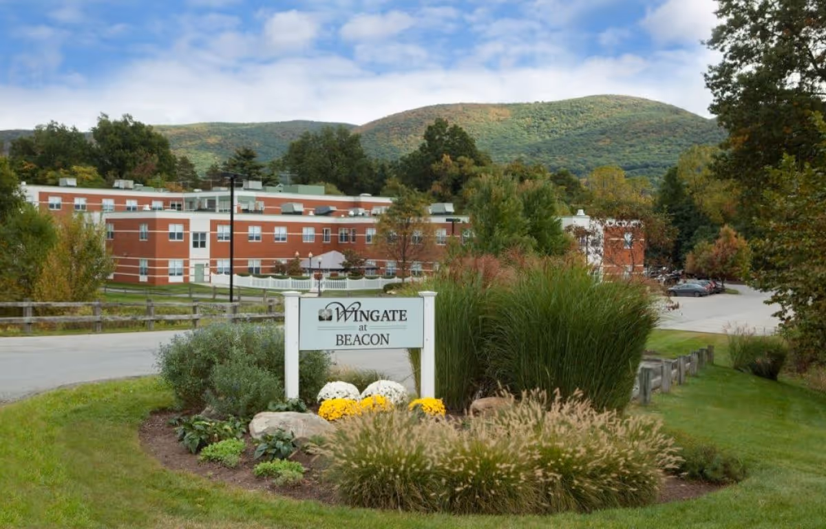 Exterior view of Wingate at Beacon facility with a landscaped garden in the foreground, a sign displaying the facility name, a parking area, and green hills in the background under a partly cloudy sky.