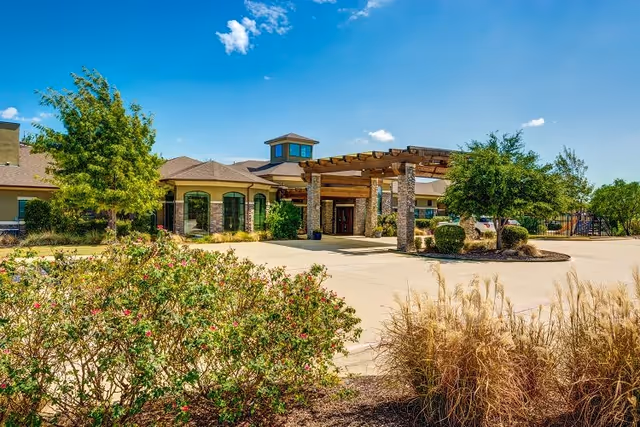 Exterior view of The Auberge at Benbrook Lake facility showing a single-story building with a covered entrance supported by stone pillars, surrounded by landscaped bushes, trees, and a clear blue sky.