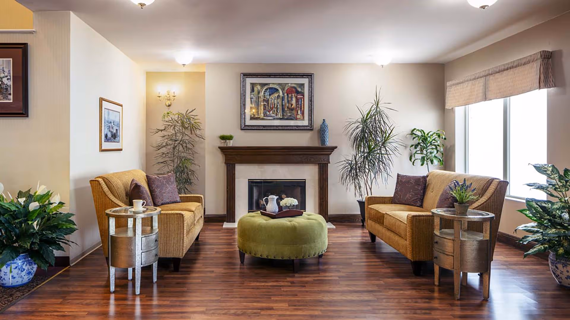 A cozy living room with two mustard yellow sofas facing each other, a green round ottoman in the center, wooden side tables with plants and a cup, a fireplace with a wooden mantel, framed artwork above the fireplace, and several potted plants around the room. The room has wooden flooring and a large window with a beige valance.