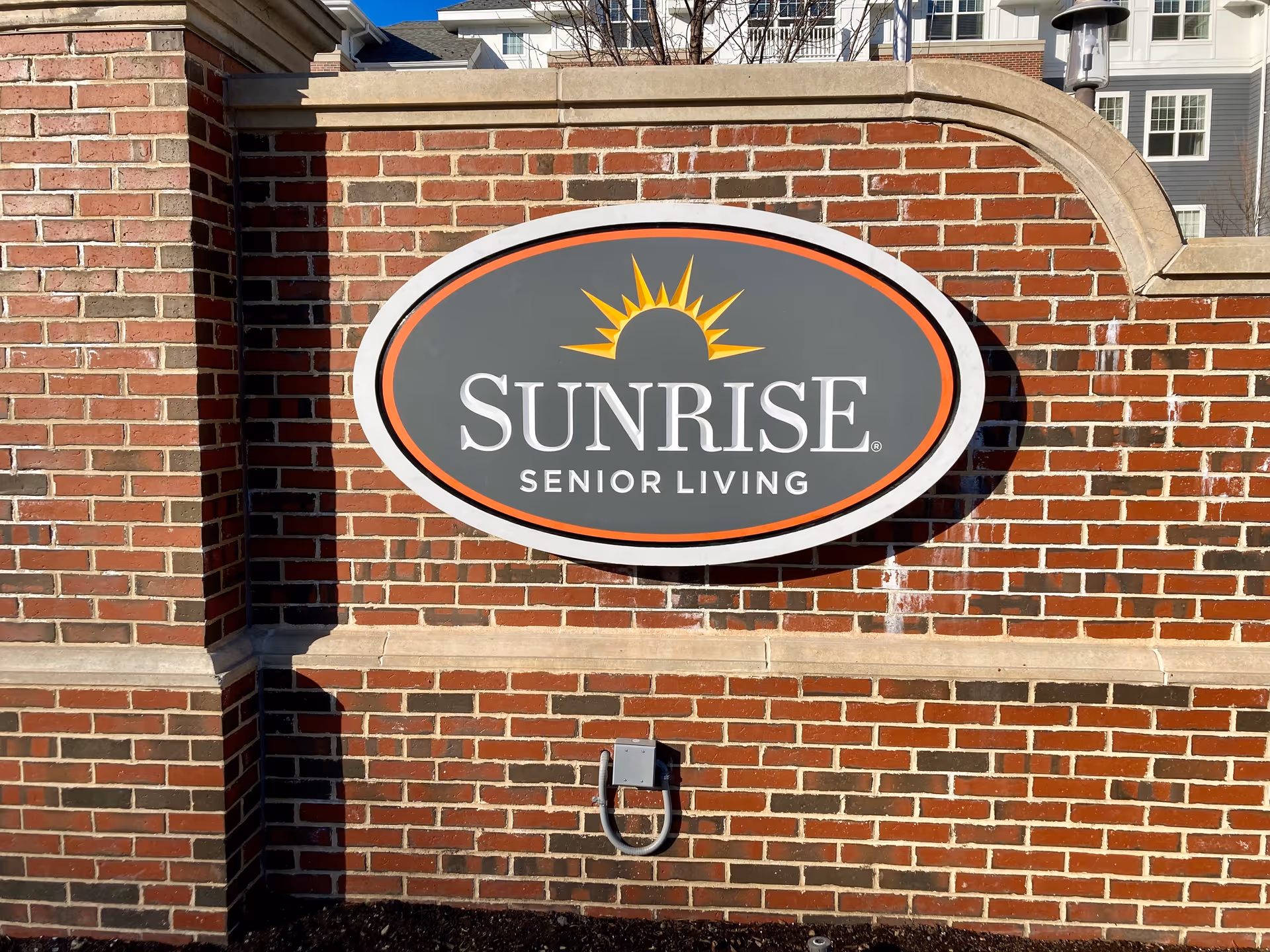 A brick wall with a large oval sign mounted on it that reads 'SUNRISE SENIOR LIVING' with a yellow sun graphic above the text.