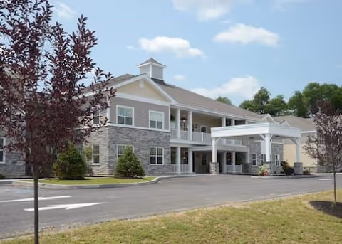 Exterior view of a two-story senior living facility building with a covered entrance, stone and siding facade, surrounded by a paved driveway, small trees, and green lawn under a partly cloudy sky.