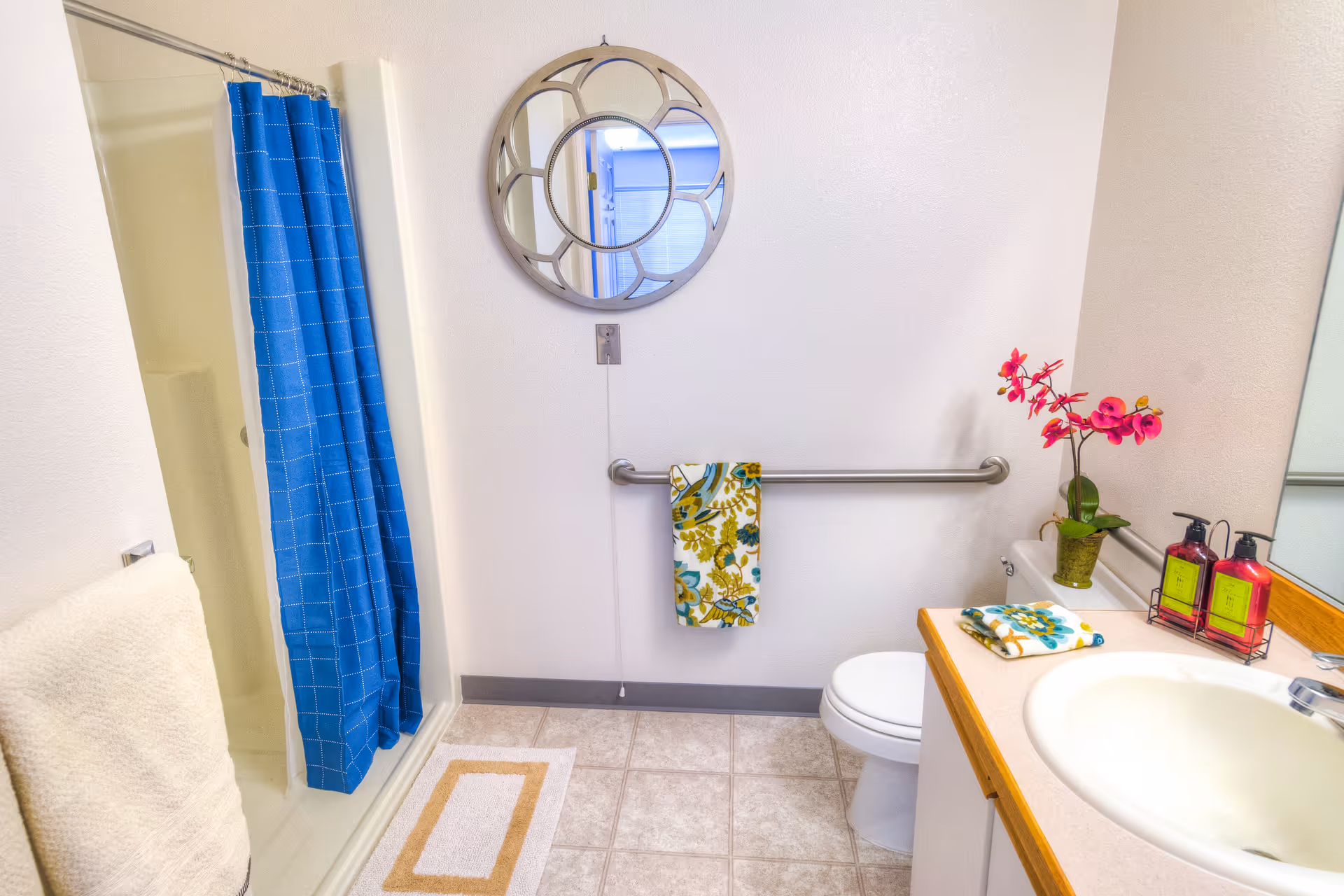 A clean and bright bathroom featuring a shower with a blue curtain, a white toilet, a sink with a wooden countertop, a round decorative mirror on the wall, a floral towel hanging on a grab bar, and a small potted pink orchid on the counter next to soap dispensers.