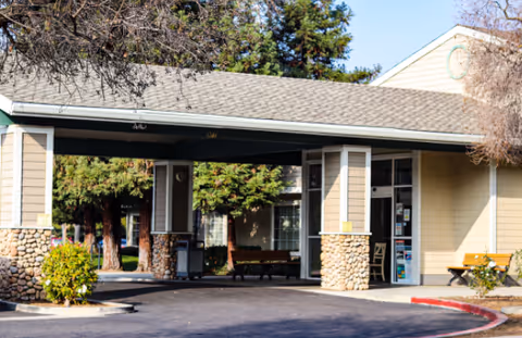 Entrance of a single-story building with a covered drop-off area supported by stone pillars, surrounded by trees and bushes under a clear sky.