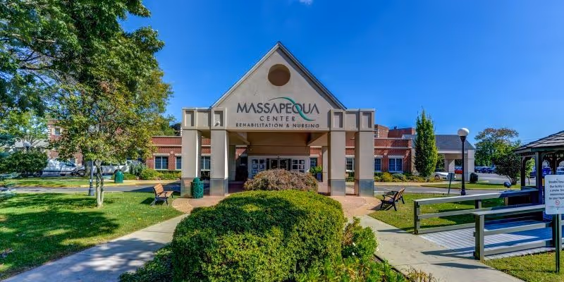 Front exterior view of Massapequa Center Rehabilitation & Nursing building with a covered entrance, surrounded by green bushes, trees, benches, and a clear blue sky.
