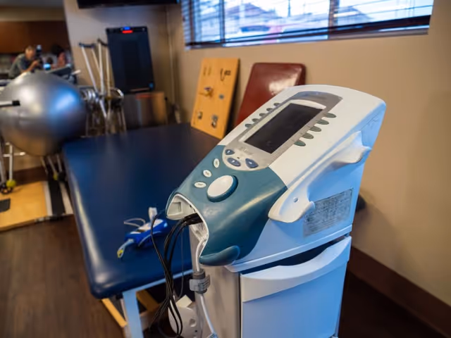 A physical therapy room with a medical device in the foreground, a blue padded therapy table, exercise equipment including a large silver exercise ball, and therapy tools in the background near a window with blinds.