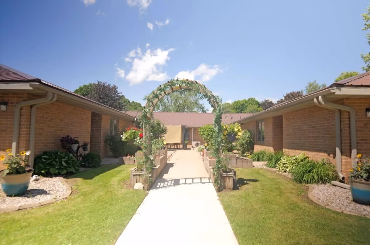 Outdoor courtyard area at Patient Pines Assisted Living featuring a paved walkway with a decorative arch covered in greenery, surrounded by green grass, flower beds, and brick buildings under a clear blue sky.