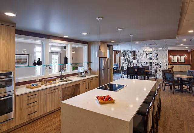 Modern kitchen area with a large white island countertop featuring a built-in stovetop and a tray of tomatoes. The kitchen has wooden cabinets, a double oven, a refrigerator, and a sink with a glass partition. In the background, there is a dining area with tables and chairs separated by decorative white panels.