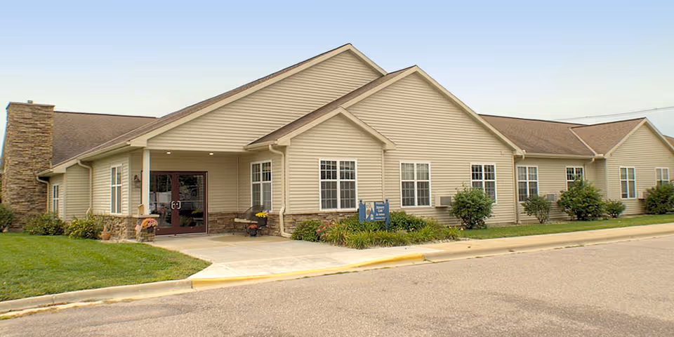 Exterior view of a single-story senior living facility building with beige siding, multiple windows, a stone chimney, and a covered entrance with double glass doors. There is a small garden with shrubs and flowers near the entrance and a paved driveway in front.