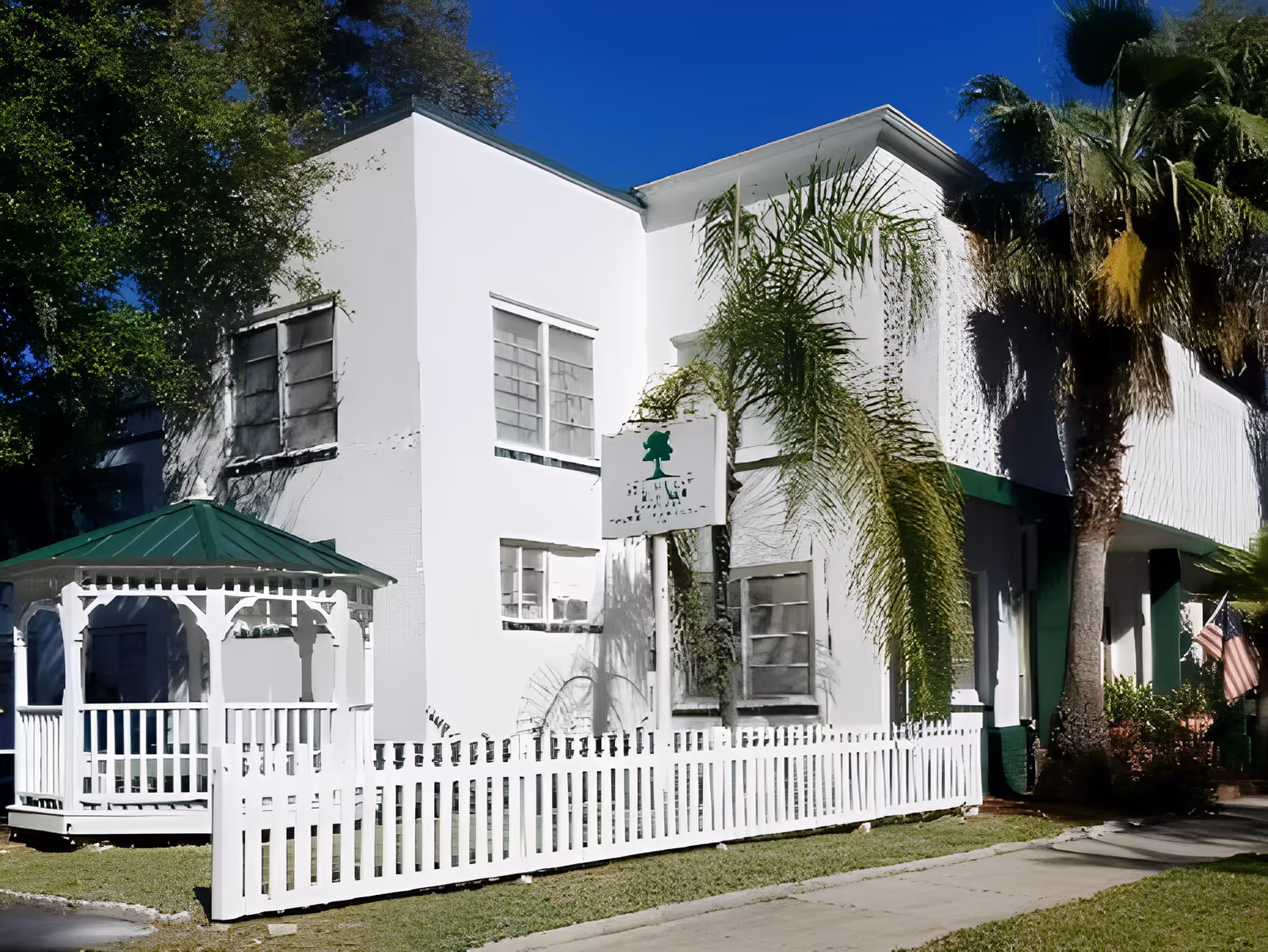 Exterior view of a white two-story building with a green roof and a white picket fence in front. There is a white gazebo with a green roof on the left side and palm trees near the building. A sign with a tree logo is mounted on the building. The sky is clear and blue.