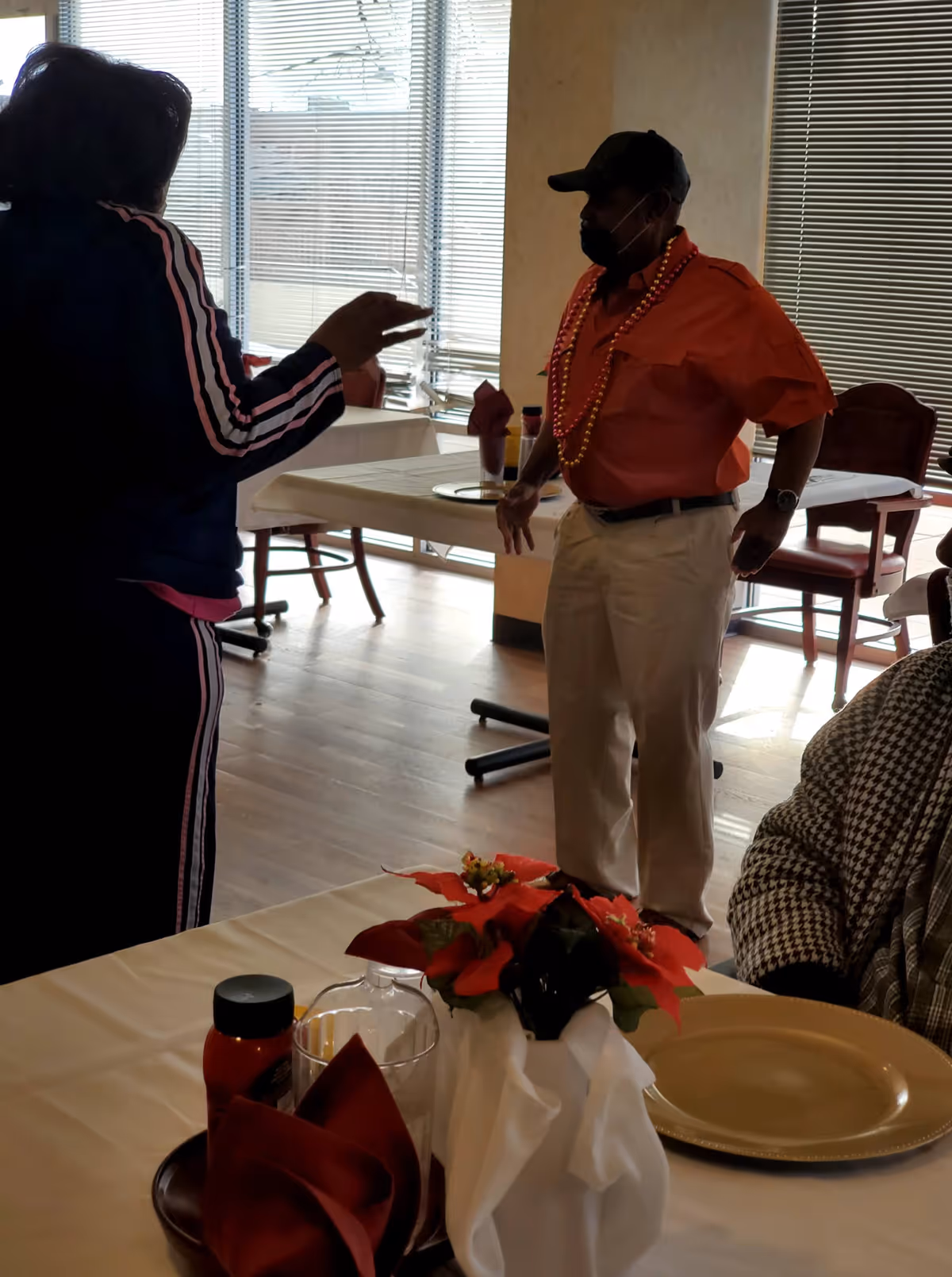 Two people standing and talking in a dining area with tables and chairs. One person is wearing a red shirt, white pants, a black cap, a face mask, and beaded necklaces. The other person is wearing a dark tracksuit with pink stripes. A table in the foreground is set with a gold plate, a glass, a bottle, and a centerpiece with red flowers and white cloth.