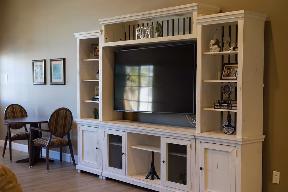 A cozy living room area featuring a large white wooden entertainment center with a flat-screen TV in the center. The shelves around the TV hold decorative items, books, and framed photos. To the left, there is a small round table with two striped upholstered chairs against a beige wall with two framed pictures.