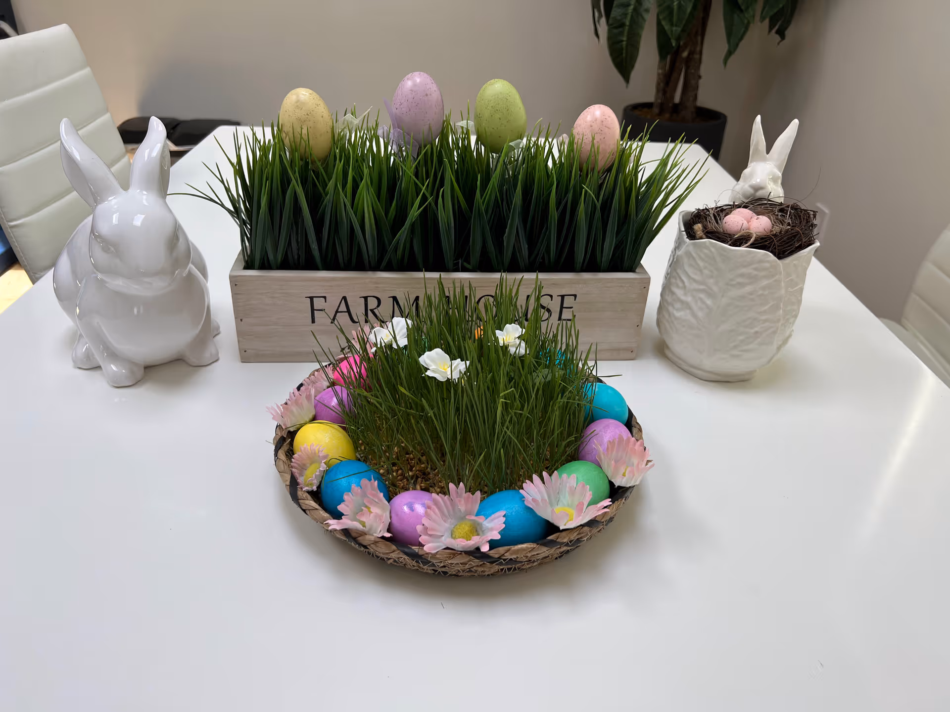 A white table with Easter-themed decorations including a white ceramic rabbit figurine, a wooden box labeled 'FARMHOUSE' filled with green grass and pastel-colored eggs, a round woven basket with grass, flowers, and colorful eggs, and a white textured pot with a small rabbit figurine and pink eggs inside.