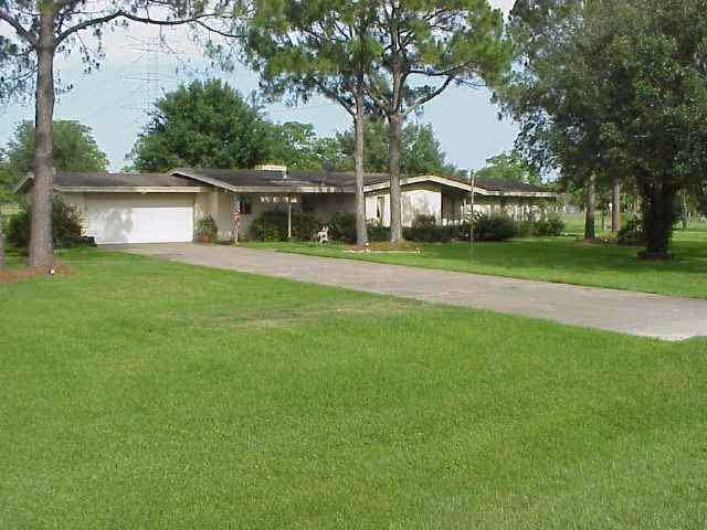Single-story residential building with a long driveway, surrounded by green grass and tall trees under a clear sky.