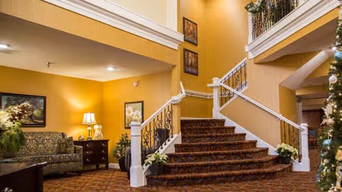 Interior view of a senior living facility lobby with a carpeted staircase featuring white railings and decorative iron balusters. To the left, there is a sitting area with a patterned sofa, a side table with a lamp, and framed artwork on the yellow walls. The space is warmly lit and decorated with plants and flowers.