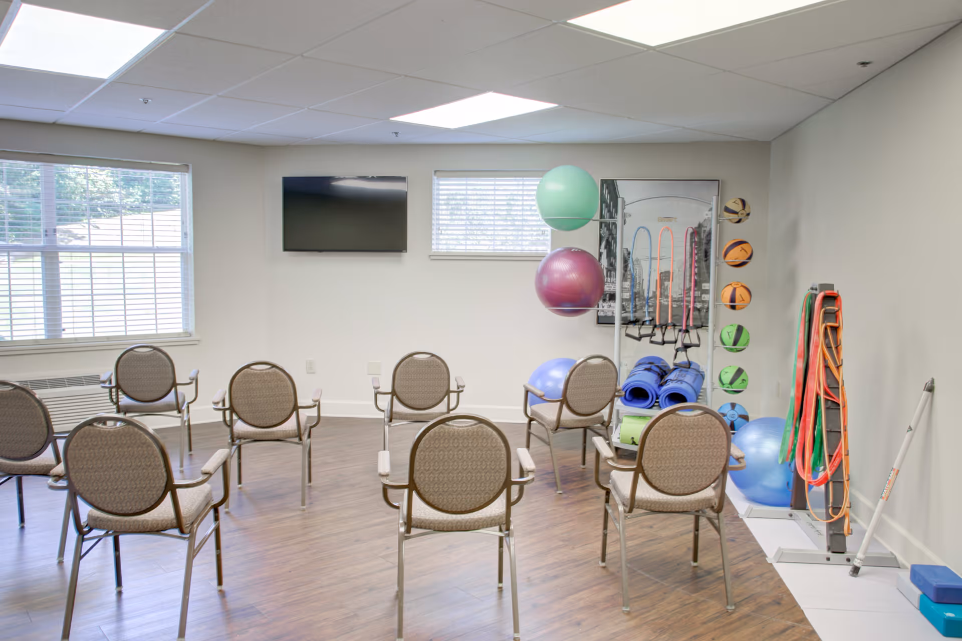A small exercise room with eight chairs arranged in two rows facing a wall-mounted TV. The room has two windows with blinds, a wooden floor, and various exercise equipment including stability balls, yoga mats, resistance bands, and small exercise balls organized on racks along one wall.