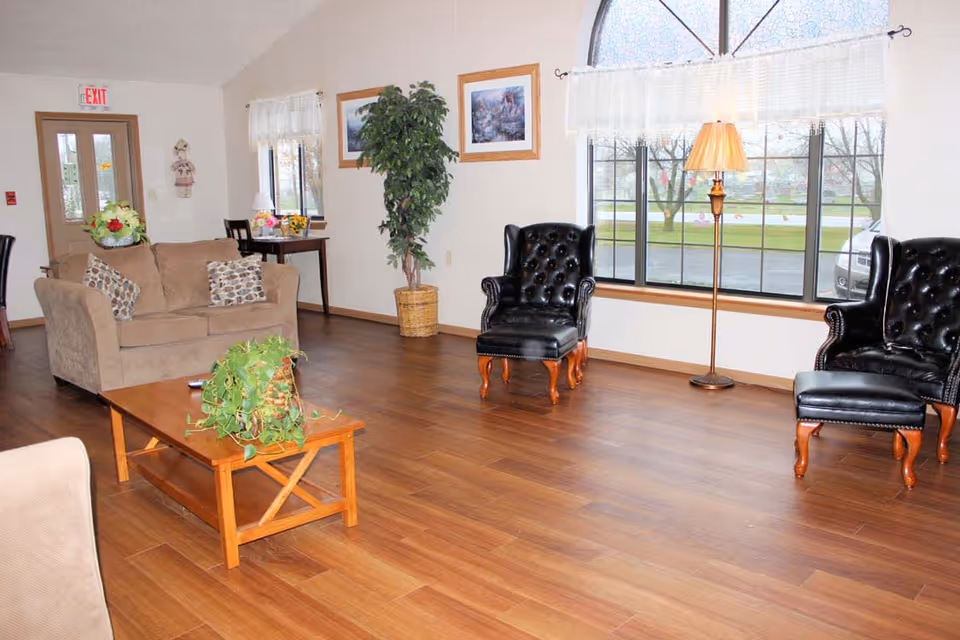 A cozy living room with wooden flooring, featuring a beige loveseat with patterned pillows, two black leather armchairs with wooden legs, a wooden coffee table with a green potted plant, a tall floor lamp, a small table with a lamp and flowers, and large windows with white curtains letting in natural light.