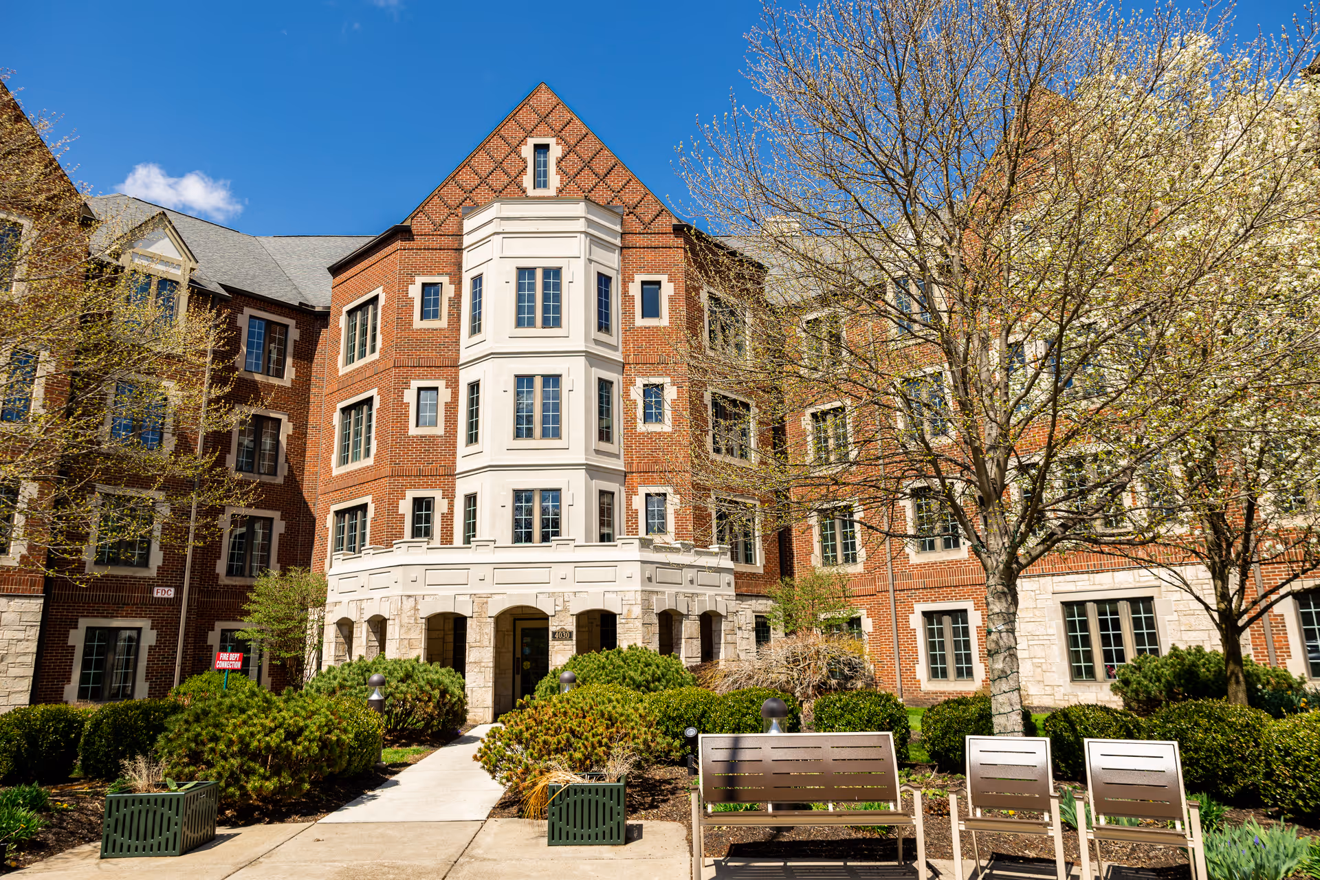 Exterior view of a multi-story senior living facility building with red brick and stone accents, surrounded by landscaped bushes and trees with early spring foliage. There are benches and a paved walkway leading to the main entrance under a clear blue sky.