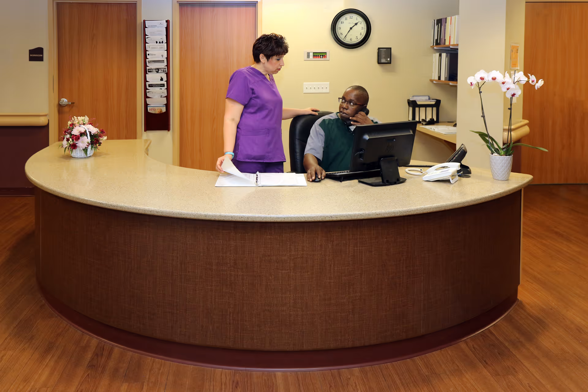 A reception area with a curved desk where a man is seated working on a computer and talking on the phone, while a woman in purple scrubs stands beside him holding papers. The background shows two wooden doors, a clock on the wall, and some office shelves with files. There are flower arrangements on the desk.