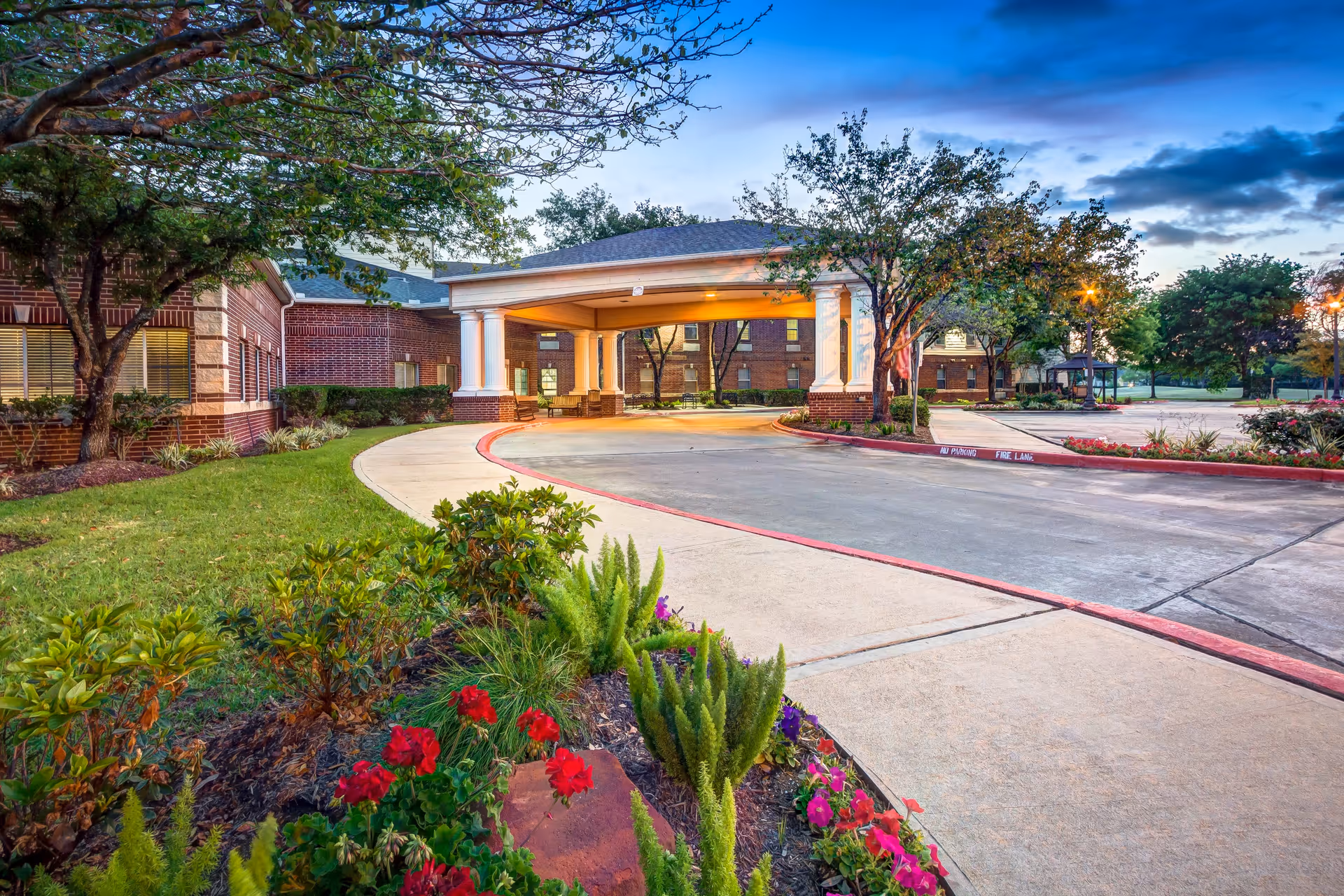 Entrance driveway to a senior living facility with a covered drop-off area supported by white columns. The building is made of red brick and surrounded by landscaped greenery, trees, and colorful flowers. The sky is partly cloudy with a hint of sunset light.