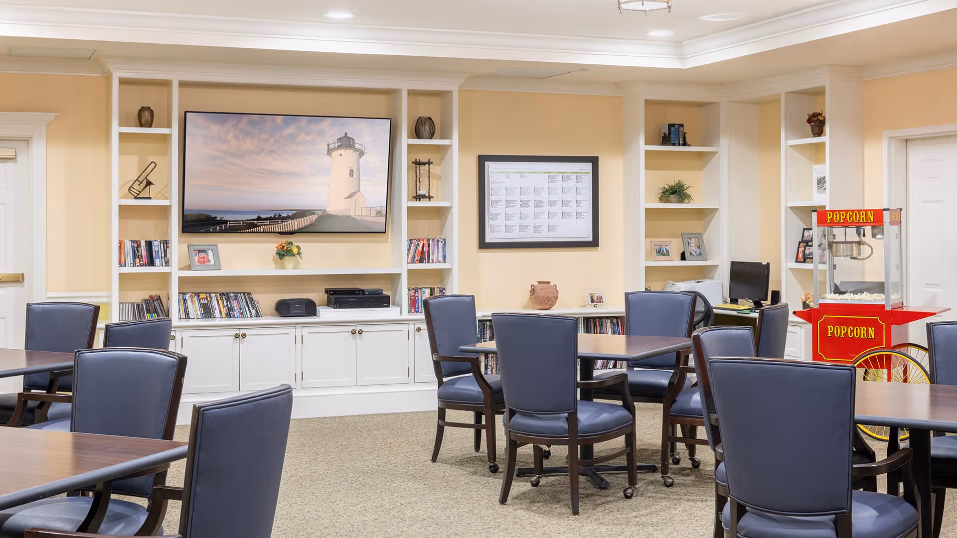 A common area in a senior living facility with multiple tables and chairs arranged for socializing or activities. The room features built-in white shelves and cabinets, a large flat-screen TV displaying a lighthouse image, a popcorn machine, and various decorative items including books, framed photos, and plants.