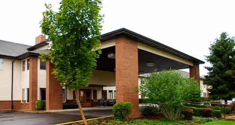 Exterior view of Brookdale McMinnville City Center showing the entrance with a covered drop-off area supported by brick columns, surrounded by greenery and trees.