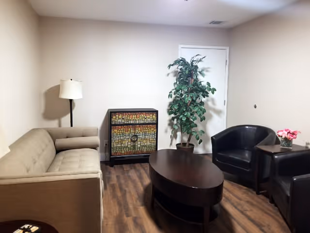 A small living room with a beige couch on the left, a floor lamp behind it, a colorful mosaic cabinet against the wall, a potted plant next to a closed white door, a dark oval coffee table in the center, and two black armchairs with a small side table holding a pink flower arrangement on the right. The room has wooden flooring and light-colored walls.