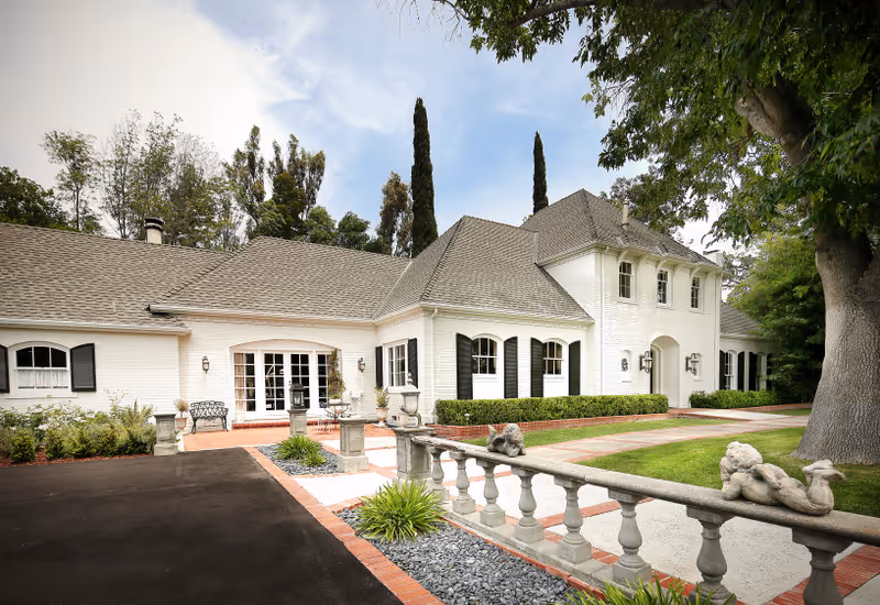 Exterior view of a large white building with a gray shingled roof, surrounded by trees and greenery. The building has multiple windows with black shutters and a paved driveway leading to the entrance. Decorative stone balustrades and small statues are visible in the foreground.