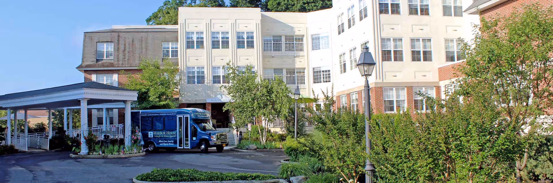 Exterior view of Harbor House Assisted Living facility showing a multi-story building with numerous windows, a covered entrance with white columns, a blue shuttle bus parked near the entrance, and landscaped greenery including trees and bushes under a clear blue sky.