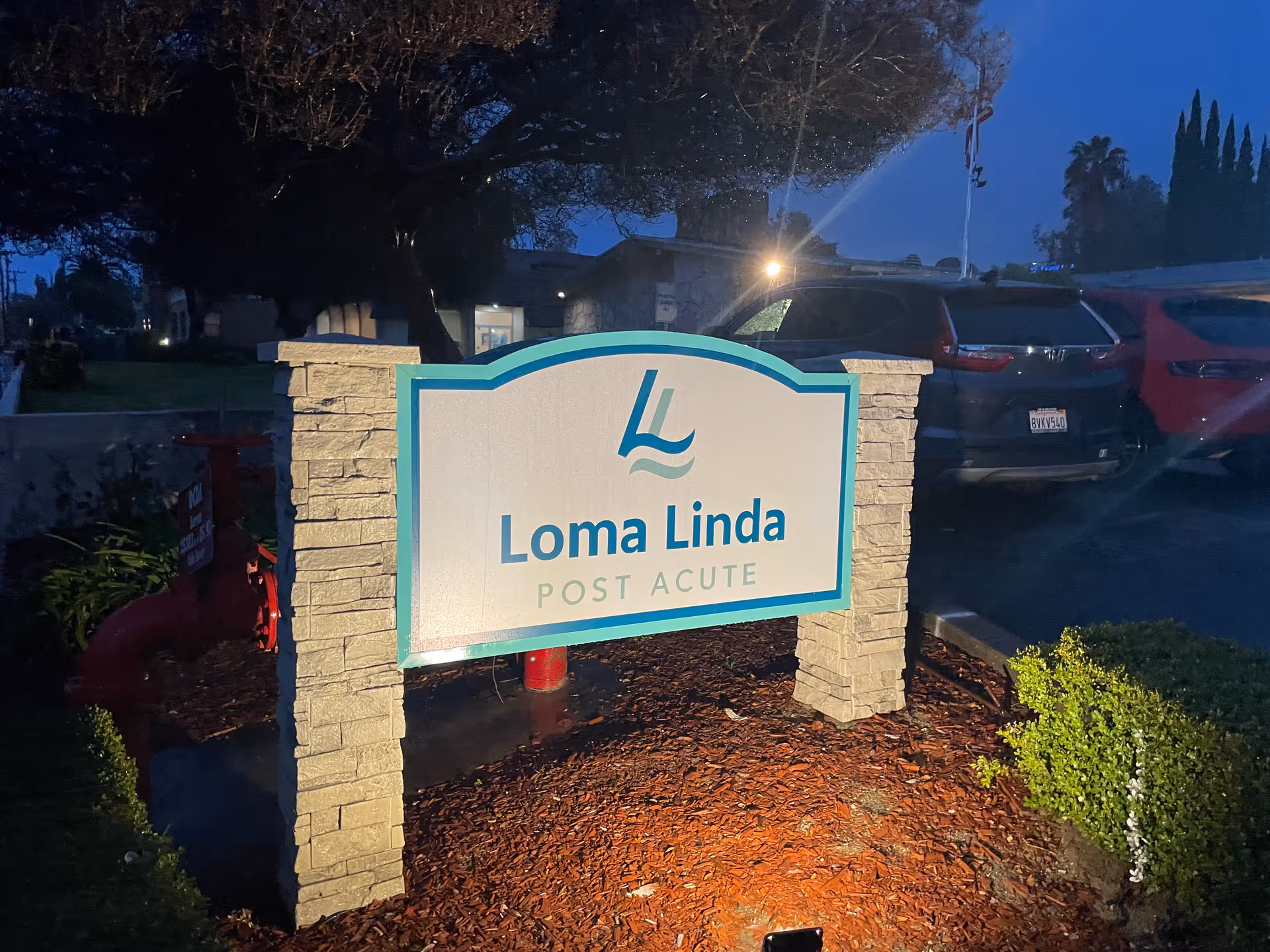 Illuminated sign for Loma Linda Post Acute facility at dusk, mounted on stone pillars with parked cars and trees in the background.