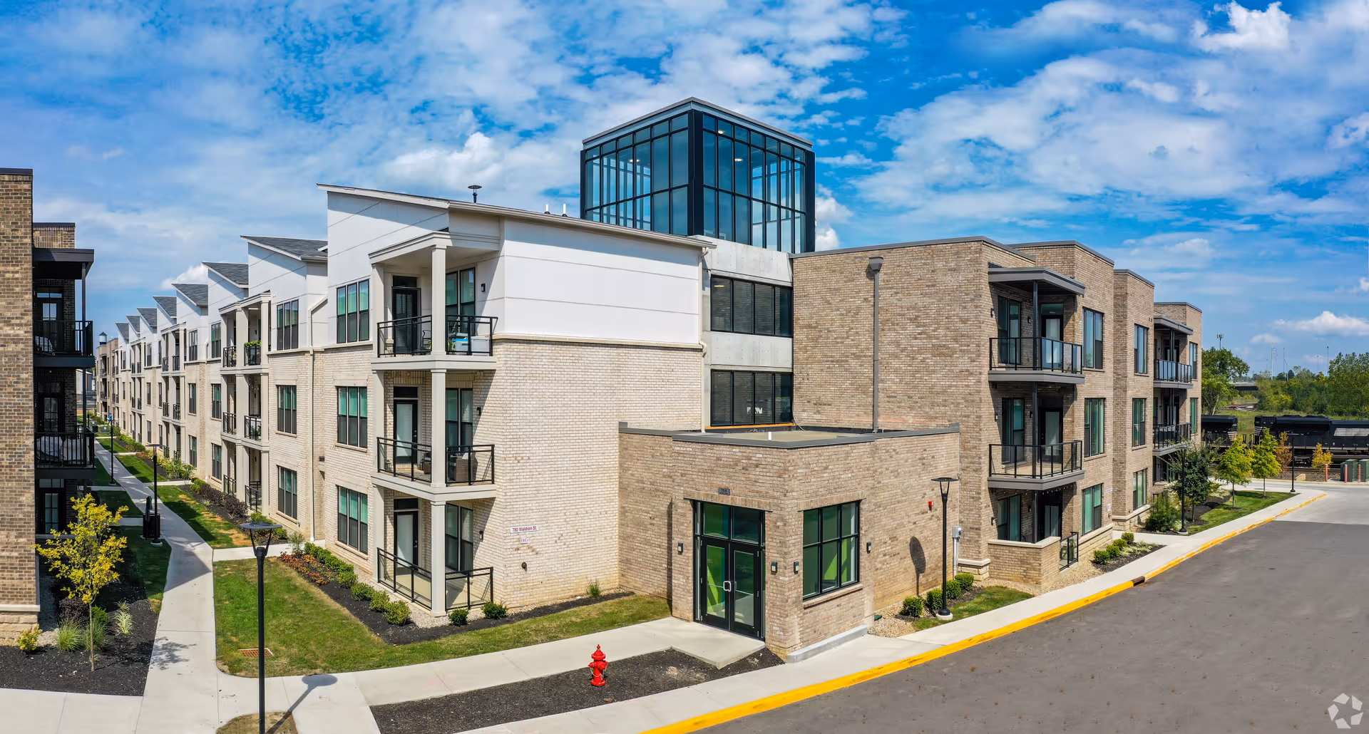 Exterior view of a modern multi-story residential building with brick and white paneling, featuring balconies and large windows under a partly cloudy blue sky.