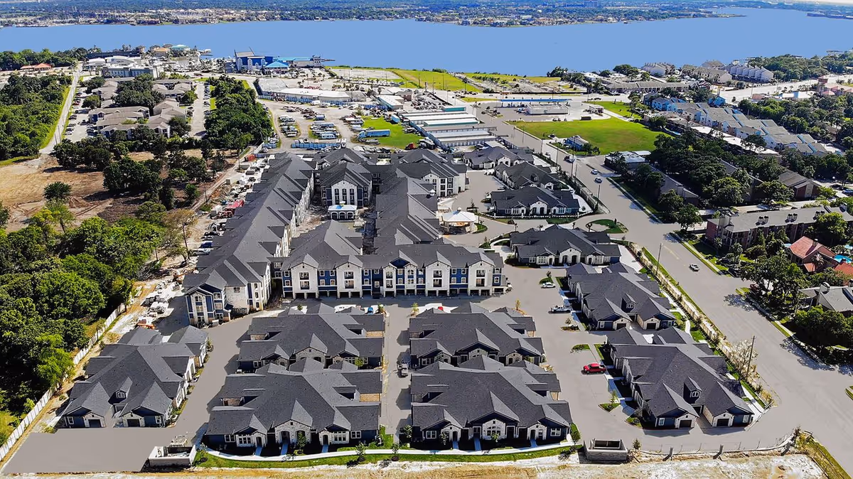 Aerial view of Chesapeake Bay Luxury Active-Adult Community showing multiple residential buildings with dark roofs arranged in a planned layout near a large body of water, surrounded by roads, greenery, and other buildings.