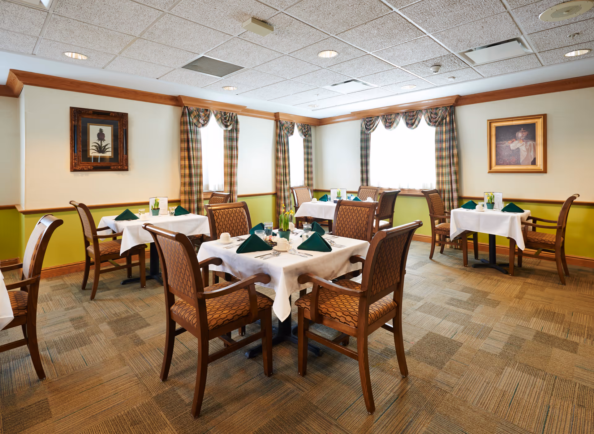 A dining room with several tables covered with white tablecloths, each set with green folded napkins, cups, and silverware. The room has patterned carpet flooring, wooden chairs with cushioned seats, plaid curtains on the windows, and framed artwork on the walls.