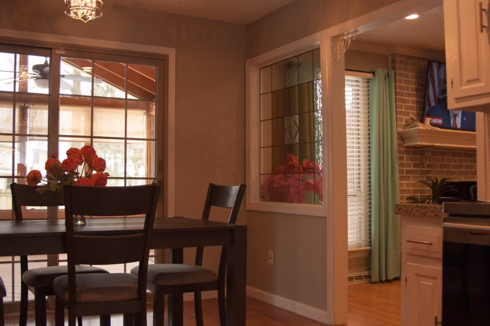 A dining area with a dark wooden table and four chairs, a vase with red flowers on the table, large sliding glass doors leading to an outdoor patio, and a window with decorative glass panels. Adjacent to the dining area is a kitchen with white cabinets, granite countertops, and a glimpse of a living room with a brick fireplace and a TV.