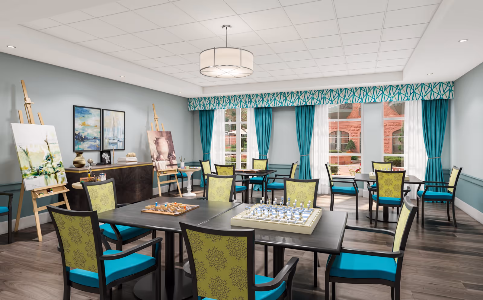 A bright and spacious activity room with multiple tables and chairs. The tables have chess and checkers boards set up for games. The room features large windows with teal curtains and valances, allowing natural light to fill the space. There are easels with paintings and art supplies along one wall, and decorative artwork hangs above a sideboard. The floor is wood, and the ceiling has recessed lighting and a central hanging light fixture.