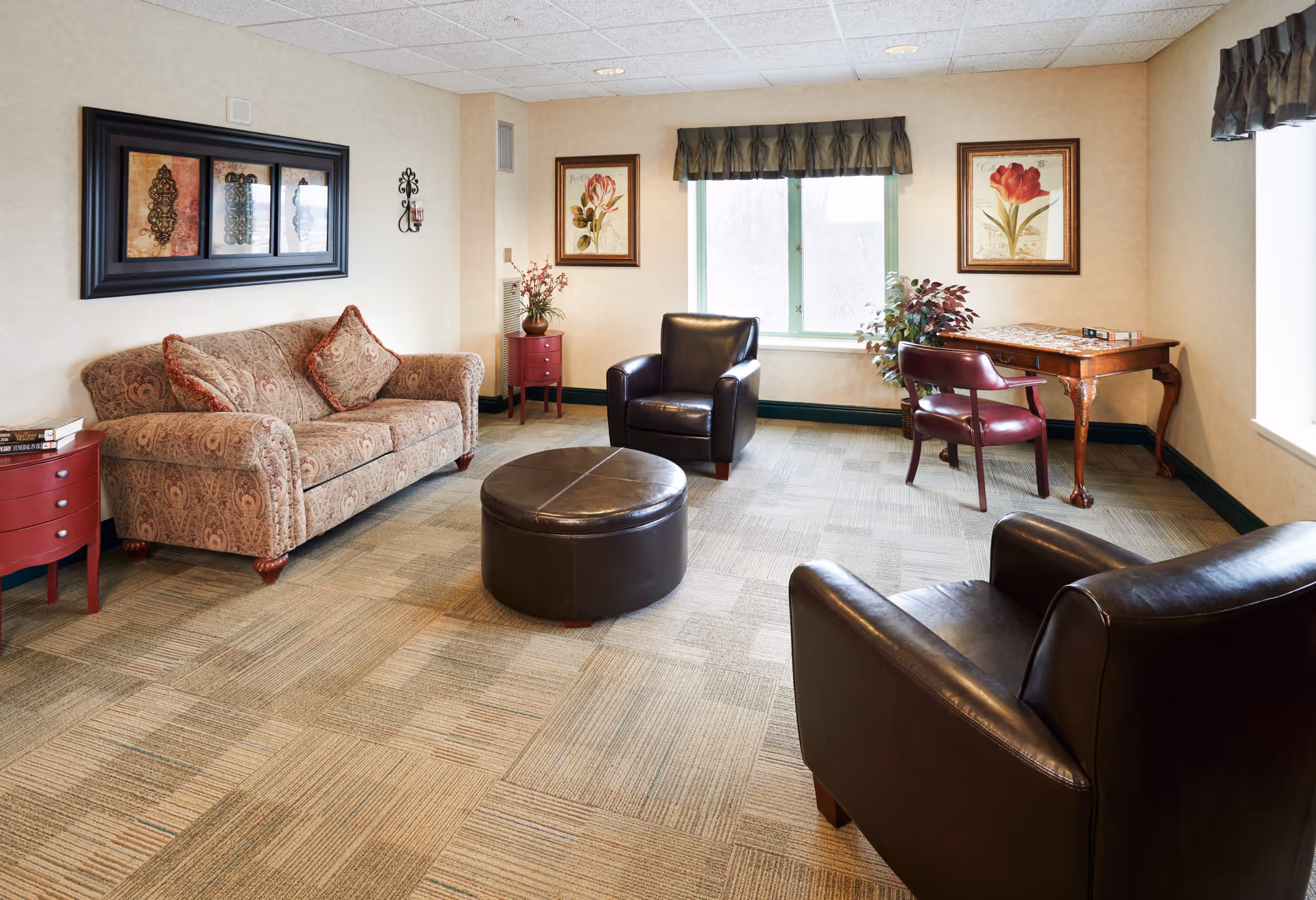 A cozy living room with a patterned sofa, two dark leather armchairs, a round leather ottoman, a wooden side table with books, a small red cabinet with a plant, a wooden desk with a chair, two framed floral paintings on the walls, and two windows with valances letting in natural light.